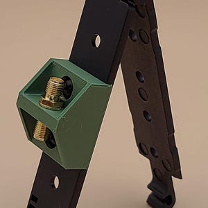 May include: A black folding mount featuring a matte olive green 3D-printed bracket attached to the center. The bracket holds a gold-colored metal SMA connector. The black frame has multiple circular mounting holes and a hinged top section.