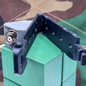May include: A black plastic hinged bracket with circular cutouts and a grey 3D-printed corner mount featuring a silver BNC connector. The component is constructed with a rigid frame and a locking mechanism, designed for mounting electronic hardware.