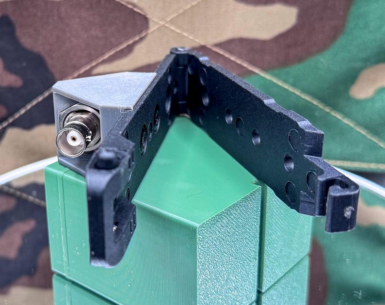 May include: A black plastic hinged bracket with circular cutouts and a grey 3D-printed corner mount featuring a silver BNC connector. The component is constructed with a rigid frame and a locking mechanism, designed for mounting electronic hardware.