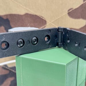 May include: A black plastic hinge component featuring a two-part articulated design with a central pivot point. The structure includes multiple circular mounting holes and integrated rivets, with a rectangular latch mechanism on one end.