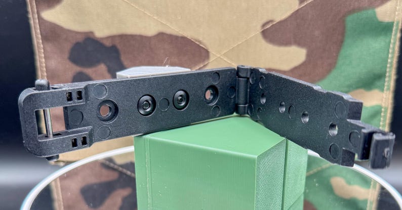 May include: A black plastic hinge component featuring a two-part articulated design with a central pivot point. The structure includes multiple circular mounting holes and integrated rivets, with a rectangular latch mechanism on one end.