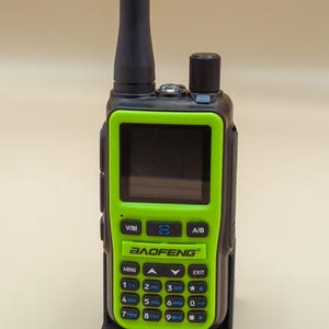 Peut inclure: Une radio bidirectionnelle portable noire et vert néon avec une longue antenne, un écran d'affichage numérique et un clavier numérique. L'appareil porte la marque BAOFENG imprimée en noir sur la façade verte, avec divers boutons de fonction.