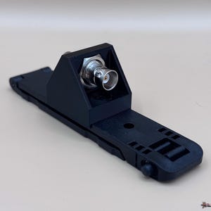 May include: A black plastic mounting bracket featuring a central BNC connector with a metallic finish. The base is elongated and rectangular with integrated slots and a small circular protrusion at one end, designed for secure equipment attachment.
