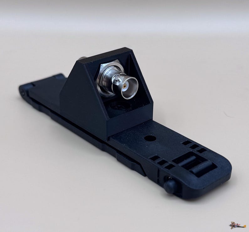 May include: A black plastic mounting bracket featuring a central BNC connector with a metallic finish. The base is elongated and rectangular with integrated slots and a small circular protrusion at one end, designed for secure equipment attachment.