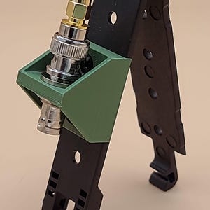 May include: A black folding mount with a green 3D-printed bracket holding a silver and gold BNC connector. The black component features multiple circular cutouts and a hinged top section, while the green bracket is secured to the frame.
