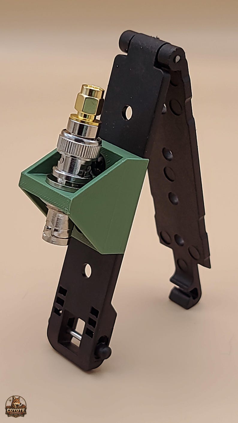 May include: A black folding mount with a green 3D-printed bracket holding a silver and gold BNC connector. The black component features multiple circular cutouts and a hinged top section, while the green bracket is secured to the frame.