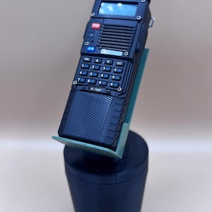 May include: A black handheld two-way radio with a digital display, a numeric keypad, and a speaker grille. The device features the text "BAOFENG" and "BF-F8HP" on the front panel. It is mounted in a teal plastic holder atop a black cylindrical base.