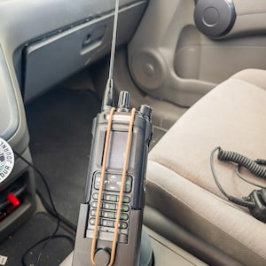 May include: A black two-way radio with an antenna, secured with a brown rubber band, sits in a car's cup holder. The radio has a keypad and a small screen. A microphone cord is visible.