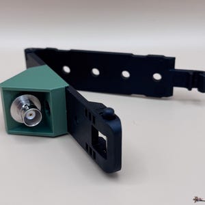 May include: A black plastic mounting bracket featuring a green triangular housing with a silver BNC connector port. The bracket includes an adjustable strap with multiple circular holes and a small protruding knob for secure attachment.