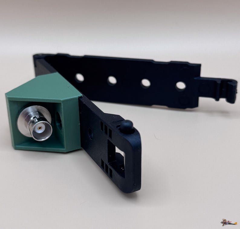 May include: A black plastic mounting bracket featuring a green triangular housing with a silver BNC connector port. The bracket includes an adjustable strap with multiple circular holes and a small protruding knob for secure attachment.