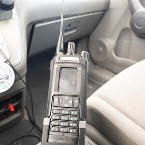 Puede incluir: Una radio port&aacute;til negra con una antena plateada est&aacute; montada en un veh&iacute;culo. La radio tiene un teclado, una pantalla y el texto "DM32" en la parte delantera. El interior del veh&iacute;culo es gris.
