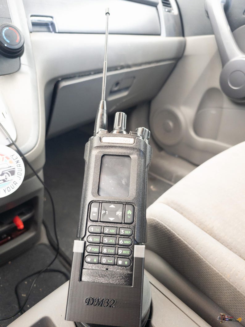Puede incluir: Una radio port&aacute;til negra con una antena plateada est&aacute; montada en un veh&iacute;culo. La radio tiene un teclado, una pantalla y el texto "DM32" en la parte delantera. El interior del veh&iacute;culo es gris.