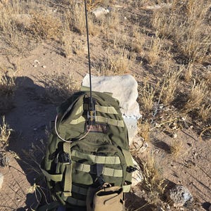 May include: An olive green tactical backpack with a long black antenna mounted on the front. The backpack features multiple nylon webbing straps, a CamelBak logo patch, and a tan glove hanging from the bottom. The design includes various buckles.