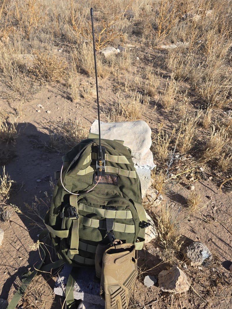May include: An olive green tactical backpack with a long black antenna mounted on the front. The backpack features multiple nylon webbing straps, a CamelBak logo patch, and a tan glove hanging from the bottom. The design includes various buckles.