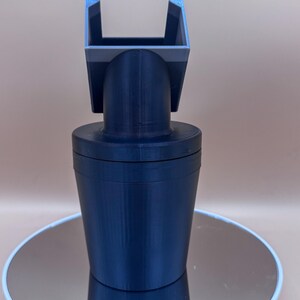Puede incluir: Un adaptador de portavasos para coche impreso en 3D de color azul oscuro. El adaptador tiene una base cil&iacute;ndrica, un cuello curvo y una parte superior cuadrada con una abertura en forma de U. El objeto est&aacute; sobre una superficie reflectante.