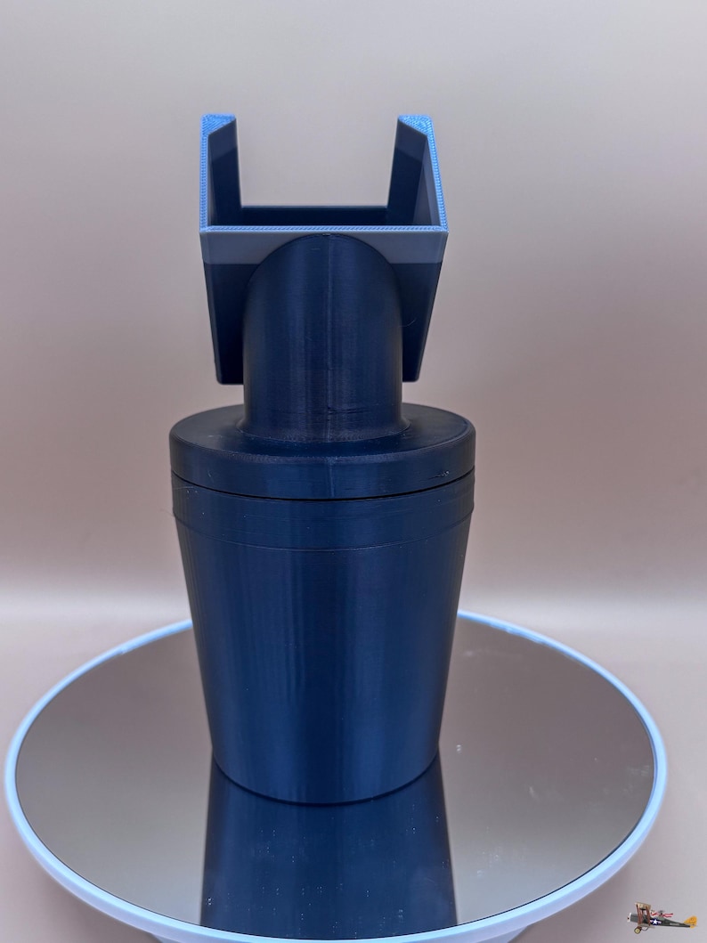 Puede incluir: Un adaptador de portavasos para coche impreso en 3D de color azul oscuro. El adaptador tiene una base cil&iacute;ndrica, un cuello curvo y una parte superior cuadrada con una abertura en forma de U. El objeto est&aacute; sobre una superficie reflectante.