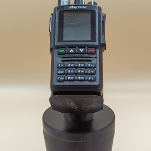 May include: A black AnyTone handheld radio with a vertical antenna, a digital display screen, and a numeric keypad featuring alphanumeric buttons. The radio is mounted in a black, cylindrical, 3D-printed plastic stand base.