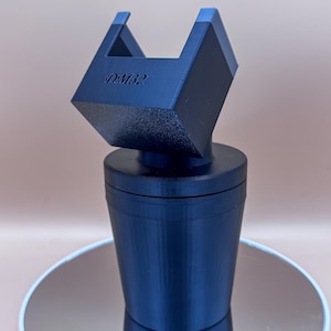 Puede incluir: Un objeto azul oscuro, impreso en 3D, con un dise&ntilde;o geom&eacute;trico. El objeto tiene una base cil&iacute;ndrica y una parte superior cuadrada con el texto "DM32". La parte superior tiene dos acentos azules. El objeto est&aacute; sobre una superficie reflectante.