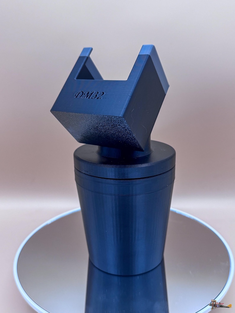 Puede incluir: Un objeto azul oscuro, impreso en 3D, con un dise&ntilde;o geom&eacute;trico. El objeto tiene una base cil&iacute;ndrica y una parte superior cuadrada con el texto "DM32". La parte superior tiene dos acentos azules. El objeto est&aacute; sobre una superficie reflectante.