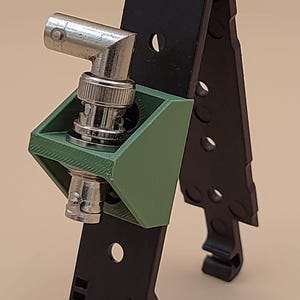 May include: A black plastic MOLLE-compatible mounting bracket featuring a green 3D-printed adapter housing a right-angle BNC connector. The black mounting strip includes multiple circular holes and a bottom locking mechanism for secure attachment.