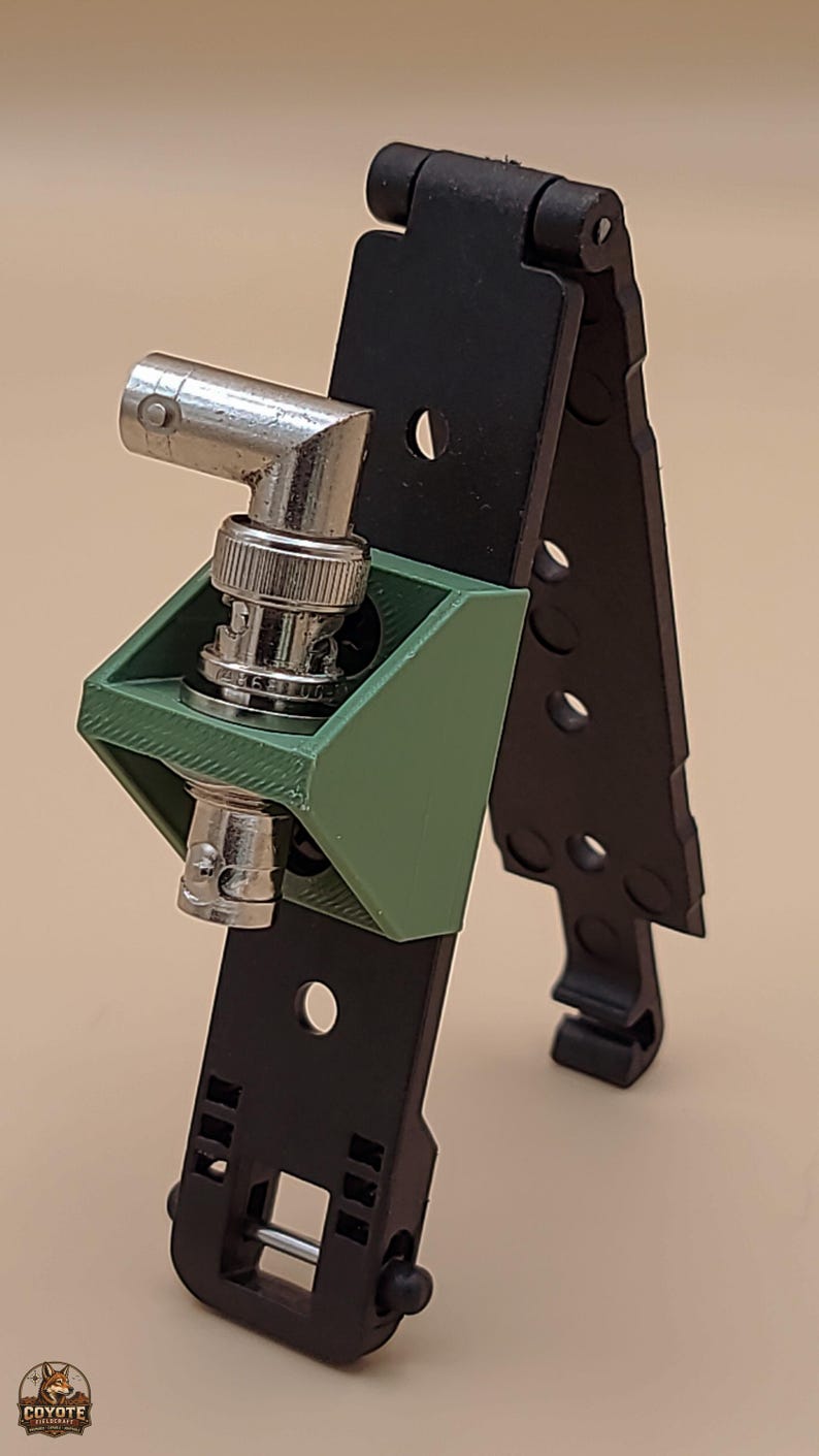 May include: A black plastic MOLLE-compatible mounting bracket featuring a green 3D-printed adapter housing a right-angle BNC connector. The black mounting strip includes multiple circular holes and a bottom locking mechanism for secure attachment.