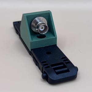 May include: A green 3D-printed mounting bracket featuring a silver BNC connector attached to a black plastic base. The base has a rectangular shape with integrated slots and a circular hole, while the bracket has a sloped front face.