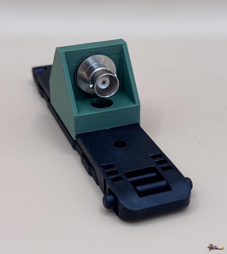 May include: A green 3D-printed mounting bracket featuring a silver BNC connector attached to a black plastic base. The base has a rectangular shape with integrated slots and a circular hole, while the bracket has a sloped front face.