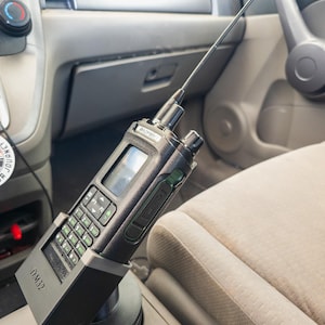 Puede incluir: Una radio port&aacute;til negra con una antena larga est&aacute; montada en el portavasos de un coche. La radio tiene un teclado, una pantalla y el texto "DM32" en la base. El interior del coche es visible, con un asiento beige y un salpicadero gris.