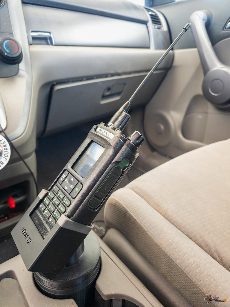 Puede incluir: Una radio port&aacute;til negra con una antena larga est&aacute; montada en el portavasos de un coche. La radio tiene un teclado, una pantalla y el texto "DM32" en la base. El interior del coche es visible, con un asiento beige y un salpicadero gris.
