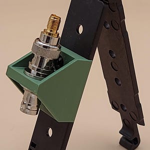 May include: A black folding antenna mount with a green 3D-printed bracket holding a silver and gold BNC connector. The mount features a hinged design with multiple circular cutouts and a small logo at the bottom left reading COYOTE FIELDCRAFT.
