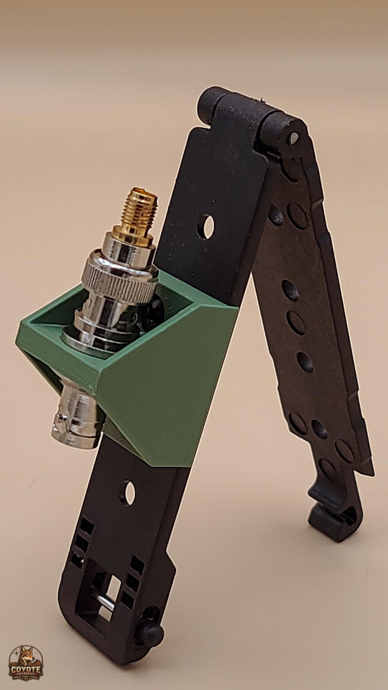 May include: A black folding antenna mount with a green 3D-printed bracket holding a silver and gold BNC connector. The mount features a hinged design with multiple circular cutouts and a small logo at the bottom left reading COYOTE FIELDCRAFT.