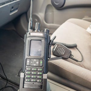 May include: A black Baofeng DM32 two-way radio with an antenna, keypad, and display screen. An AnyTone microphone is connected. The radio is mounted in a vehicle's console.