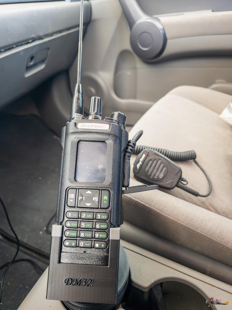 Puede incluir: Una radio bidireccional negra Baofeng DM32 con antena, teclado y pantalla. Un micr&oacute;fono AnyTone est&aacute; conectado. La radio est&aacute; montada en la consola de un veh&iacute;culo.