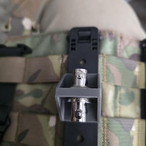 May include: A black and gray tactical accessory with a silver connector, attached to a camouflage vest. The accessory appears to be a mount for a device, with a rectangular design and a secure attachment mechanism.
