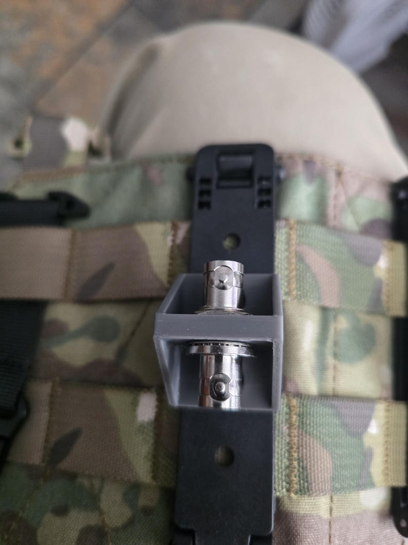 May include: A gray plastic bracket holding two silver BNC connectors, mounted onto a black vertical strap with a camouflage pattern background. The bracket features a rectangular frame design with circular openings for the metal connectors.