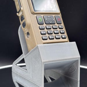 May include: A tan-colored, ruggedized mobile phone with a keypad and screen, resting in a light gray 3D-printed stand. The phone has black buttons and a screen displaying a blank interface. The stand has a geometric design.