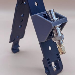 May include: A dark blue, hinged, multi-hole mounting strap with a grey, 3D-printed, triangular bracket attached. A metallic BNC connector is secured within the bracket, featuring a silver-toned cylindrical body and a gold-toned threaded base.
