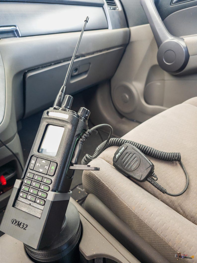Puede incluir: Una radio de dos v&iacute;as negra con pantalla, teclado y antena montada en el portavasos de un coche. La radio est&aacute; etiquetada como "DM32" y tiene un micr&oacute;fono conectado etiquetado como "AnyTone".