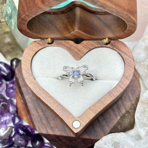 Puede incluir: Un anillo de mariposa plateado con una piedra central azul claro se encuentra dentro de una caja de madera en forma de corazón. La caja está abierta, revelando un interior blanco. El anillo presenta detalles intrincados y la caja es de madera oscura.