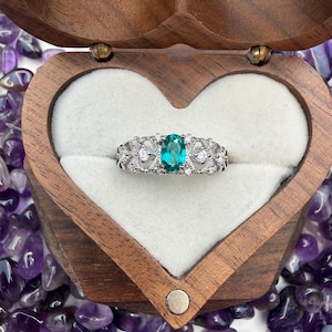 以下が含まれることがあります： 中央に楕円形のティール色の宝石、周囲に小さな白い石と装飾的な透かし模様が施されたシルバーリング。 リングは、白い内側のハート型の木製ジュエリーボックスに飾られています。