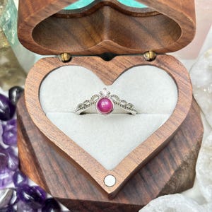 Puede incluir: Un anillo de plata con una gema rosa redonda, engastada en un diseño en forma de corona. El anillo se muestra en una caja de madera en forma de corazón con un interior blanco. La caja está abierta.