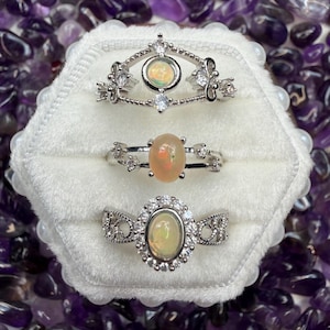 May include: Three silver rings with opal gemstones are displayed on a white velvet surface. The rings have intricate designs, including floral and crown-like elements, and are surrounded by small, sparkling crystals. The background features purple amethyst stones.
