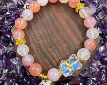 May include: A beaded bracelet featuring a mix of pink, peach, and clear round gemstones. The bracelet includes gold-colored accents and a decorative charm with a flower design. The bracelet is displayed on a wooden surface surrounded by purple stones.