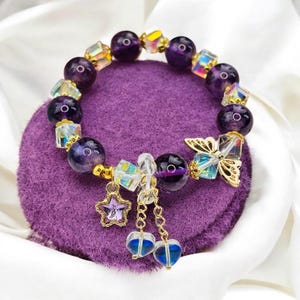 May include: A bracelet with deep purple, round beads and iridescent, square-cut crystals. Gold charms include a butterfly, a star, and two heart pendants. The bracelet is displayed on a purple velvet surface.
