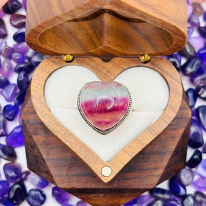 Puede incluir: Un anillo en forma de corazón con una piedra preciosa rosa, verde y blanca engastada en una banda de plata. El anillo se presenta en una caja de madera en forma de corazón.