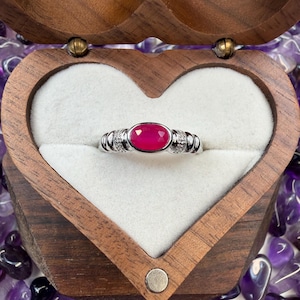 May include: A silver ring featuring a large oval ruby gemstone, set between smaller clear stones. The ring is presented in a heart-shaped wooden box with a white lining, surrounded by purple amethyst crystals.