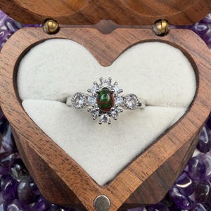 May include: A silver ring with a dark green and red oval-shaped stone at the center, surrounded by smaller white stones in a floral design. The ring is displayed in a heart-shaped wooden box with a white interior.