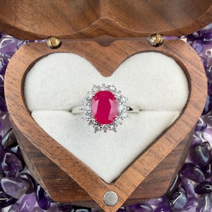May include: A silver ring with a large oval ruby center stone, surrounded by smaller white gemstones. The ring is displayed in a heart-shaped wooden box with a white interior, set against a background of purple gemstones.