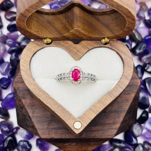 May include: A heart-shaped wooden ring box with a white velvet interior. A silver ring with a large oval-shaped ruby gemstone surrounded by smaller diamonds is inside the box.