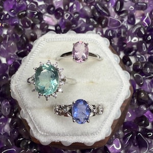 Peut inclure: Trois bagues en pierres précieuses présentées dans un coffret en bois à intérieur blanc, sur un fond de pierres d'améthyste violettes. Les bagues présentent des pierres ovales roses clair, bleu-vert et bleues, avec des montures en argent.